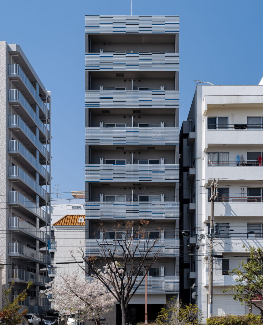 両側の建物に挟まれて建つ集合住宅。前景に桜の木や植栽が見える街中の風景