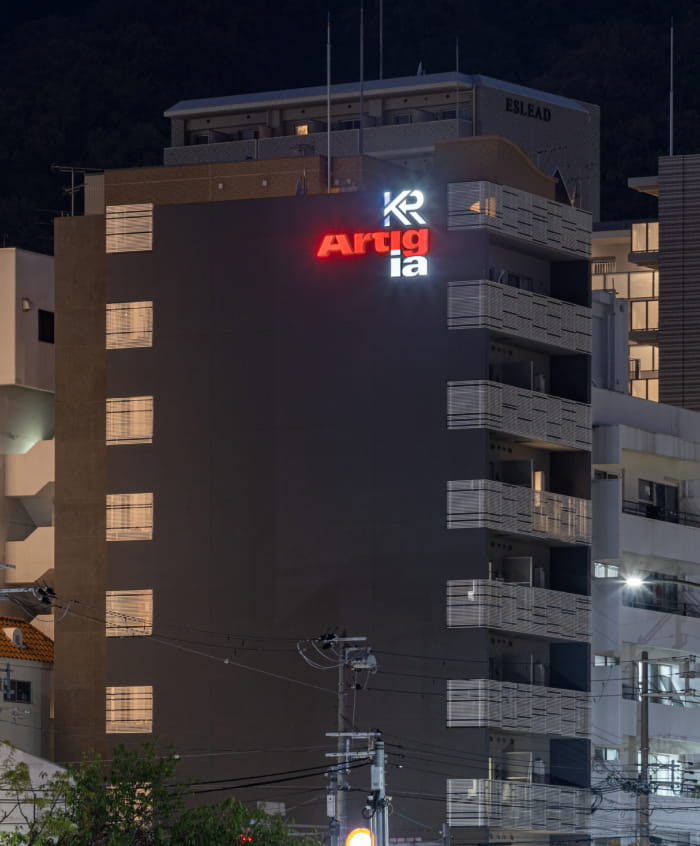 バルコニーが並ぶ集合住宅の夜景。建物上部のロゴサインと周囲の街灯が光る。